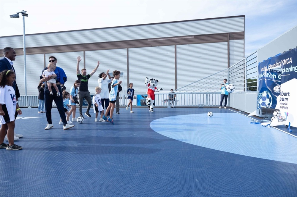 adults and youth exclaim after the completion of the first kick on the dark blue mini-pitch. a decorative banner breaks away on the face of the goal.